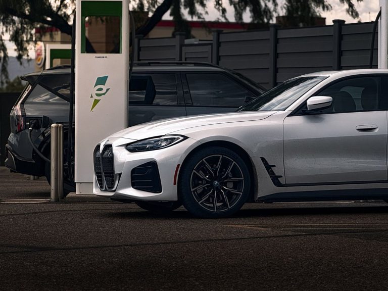 Carro elétrico branco sendo recarregado em uma estação pública, com outro veículo ao lado.